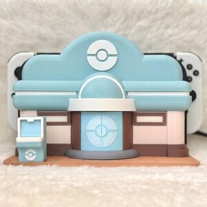 May include: A light blue and brown decorative Nintendo Switch charging station shaped like a building. The building has a light blue roof with a white and blue logo. The base is brown, and the walls are pink and brown.