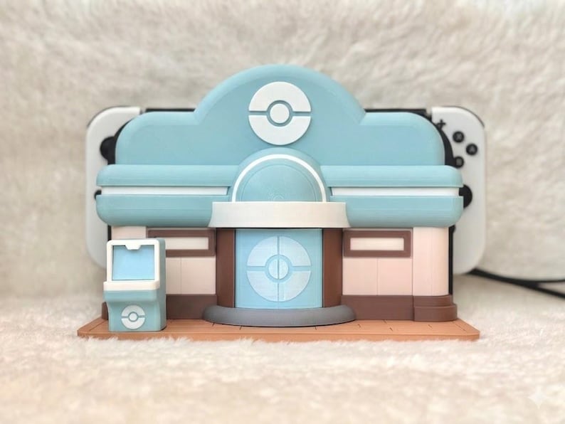 May include: A light blue and brown decorative Nintendo Switch charging station shaped like a building. The building has a light blue roof with a white and blue logo. The base is brown, and the walls are pink and brown.