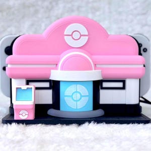 May include: A pink and white Nintendo Switch dock cover designed to resemble a Pok&eacute;mon Center building. The cover features a central blue door with a white Pok&eacute; Ball emblem, a matching pink roof with a Pok&eacute; Ball icon, and a small pink kiosk.
