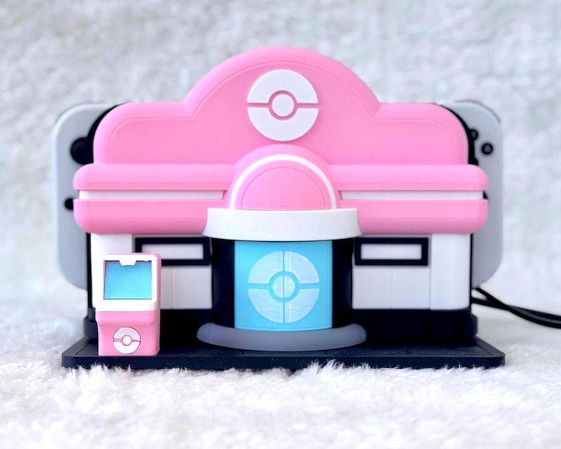 May include: A pink and white Nintendo Switch dock cover designed to resemble a Pok&eacute;mon Center building. The cover features a central blue door with a white Pok&eacute; Ball emblem, a matching pink roof with a Pok&eacute; Ball icon, and a small pink kiosk.