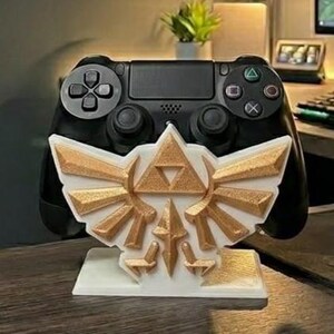 May include: A black PlayStation 4 controller resting on a white and gold-colored stand. The stand features a stylized design with a triangle and wing motif. The controller is in focus, with the stand providing support.
