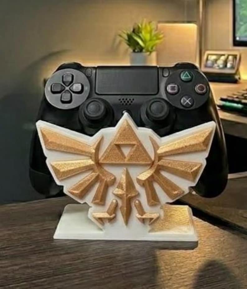 May include: A black PlayStation 4 controller resting on a white and gold-colored stand. The stand features a stylized design with a triangle and wing motif. The controller is in focus, with the stand providing support.