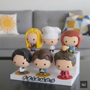 May include: A white display stand featuring seven cartoon figurines of characters from the TV show "Friends." The figurines have large heads and small bodies. The word "FRIENDS" is printed on the front of the stand.