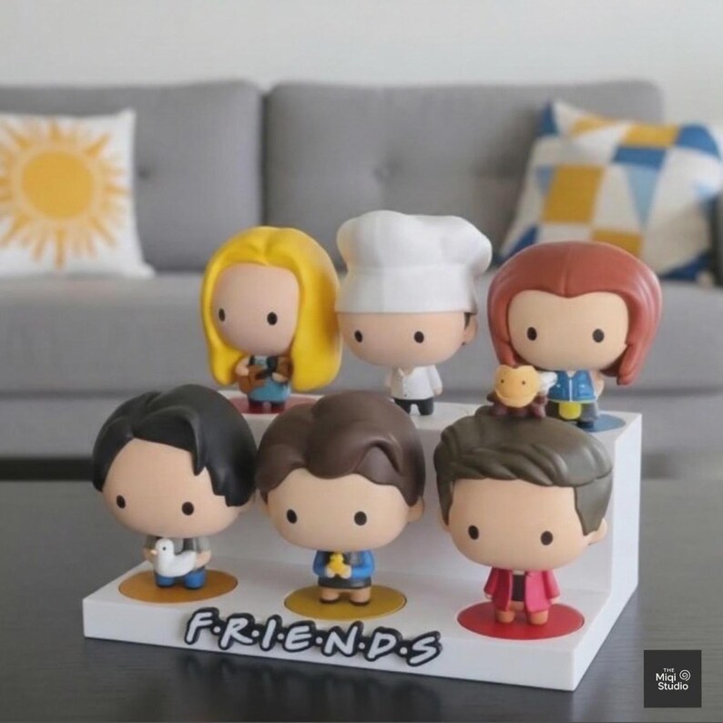 May include: A white display stand featuring seven cartoon figurines of characters from the TV show "Friends." The figurines have large heads and small bodies. The word "FRIENDS" is printed on the front of the stand.