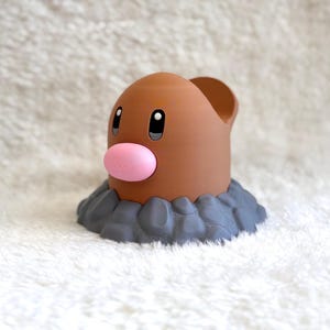 May include: A brown, cylindrical figurine with a smooth, rounded top and a prominent pink oval nose. The figure features two black eyes with white highlights and is set into a base of textured, dark gray rocks, resembling a creature emerging from earth.