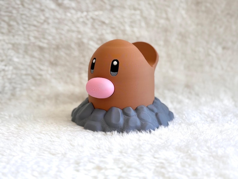 May include: A brown, cylindrical figurine with a smooth, rounded top and a prominent pink oval nose. The figure features two black eyes with white highlights and is set into a base of textured, dark gray rocks, resembling a creature emerging from earth.