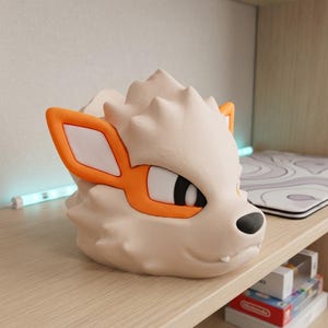 May include: A cream-colored, stylized Arcanine head figurine with orange accents around the eyes and ears, and a black nose. The figurine is sitting on a wooden shelf. The design is reminiscent of a popular animated series.