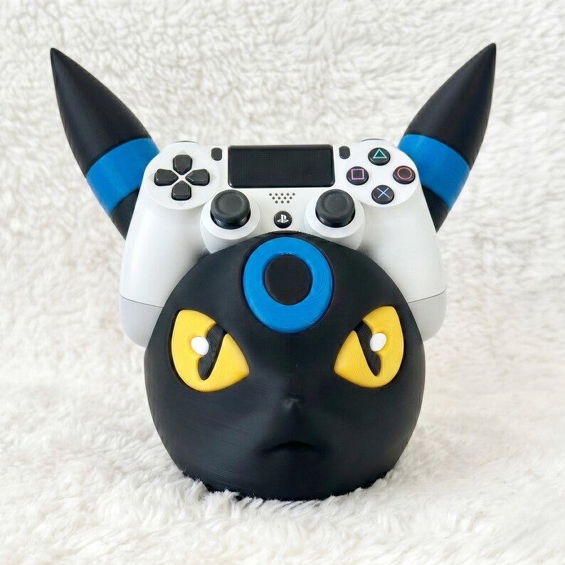 May include: A black and blue Umbreon-themed controller stand featuring yellow eyes and a blue forehead ring, holding a white PlayStation 4 game controller with black analog sticks, buttons, and a central touchpad with the PlayStation logo.