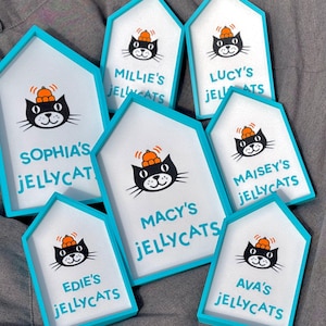 May include: A collection of house-shaped signs with a white background and a teal border. Each sign features a black cat illustration wearing a hat and the text "Jellycats" with a name above it. The signs are arranged on a gray surface.