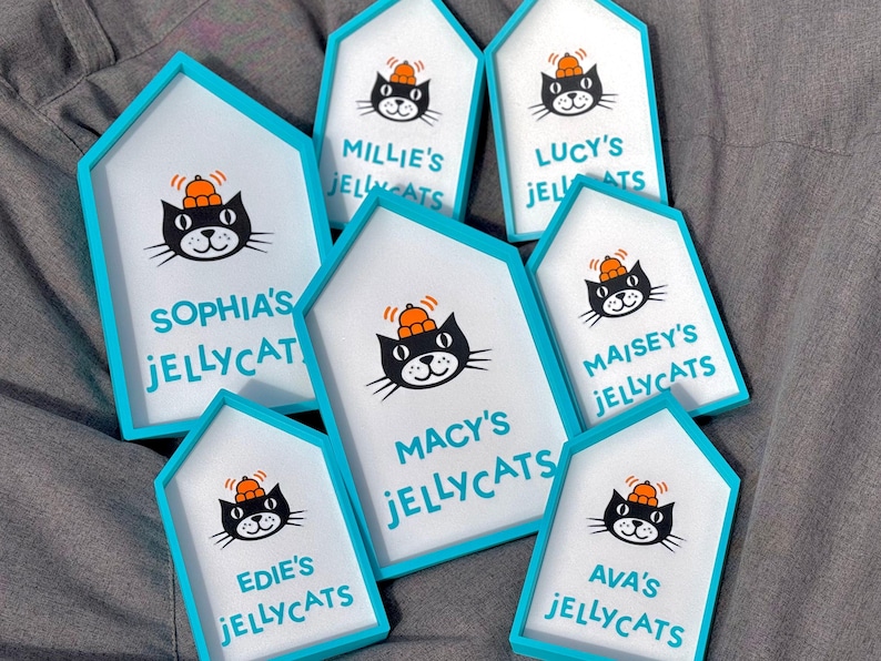 May include: A collection of house-shaped signs with a white background and a teal border. Each sign features a black cat illustration wearing a hat and the text "Jellycats" with a name above it. The signs are arranged on a gray surface.