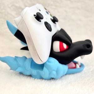 May include: A white video game controller resting on a black and light blue dragon-shaped stand. The stand features a black dragon head with red eyes and white teeth, set against a light blue cloud-like base with blue accents on the ears.