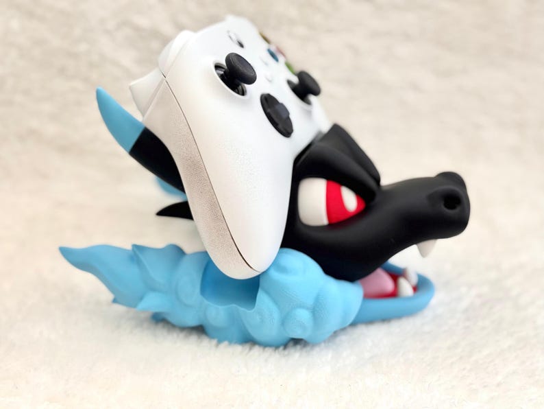 May include: A white video game controller resting on a black and light blue dragon-shaped stand. The stand features a black dragon head with red eyes and white teeth, set against a light blue cloud-like base with blue accents on the ears.