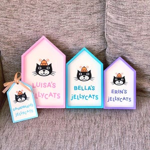 Puede incluir: Tres letreros de madera en forma de casa con marcos rosa, azul y morado, cada uno con una ilustraci&oacute;n de un gato negro con un sombrero naranja y el texto 'LUISA'S JELLYCATS', 'BELLA'S JELLYCATS' y 'ERIN'S JELLYCATS'. Una etiqueta peque&ntilde;a dice 'SAVANNAH'S JELLYCATS'.