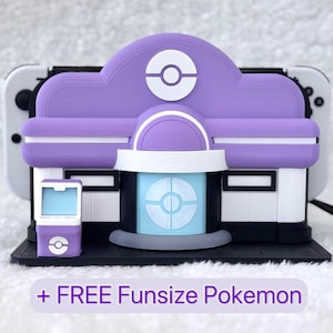 Puede incluir: Una base de carga para consola de juegos en color morado y blanco con forma de edificio, un emblema circular, una puerta azul claro y un pequeño soporte a juego. El texto + FREE Funsize Pokemon aparece en la parte inferior.