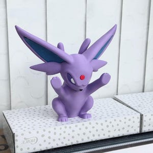 Puede incluir: Una figura de Espeon de color morado con orejas grandes y puntiagudas, detalles en azul oscuro en el interior de las orejas y una peque&ntilde;a gema circular roja en la frente. La criatura est&aacute; en una postura sentada con las patas delanteras.