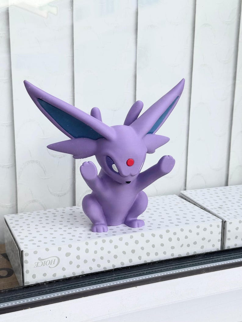 Puede incluir: Una figura de Espeon de color morado con orejas grandes y puntiagudas, detalles en azul oscuro en el interior de las orejas y una peque&ntilde;a gema circular roja en la frente. La criatura est&aacute; en una postura sentada con las patas delanteras.