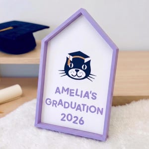 May include: A house-shaped sign with a lavender frame, featuring a cat wearing a graduation cap. The sign reads "AMELIA'S GRADUATION 2026" in lavender lettering. A graduation cap and diploma are visible in the background.