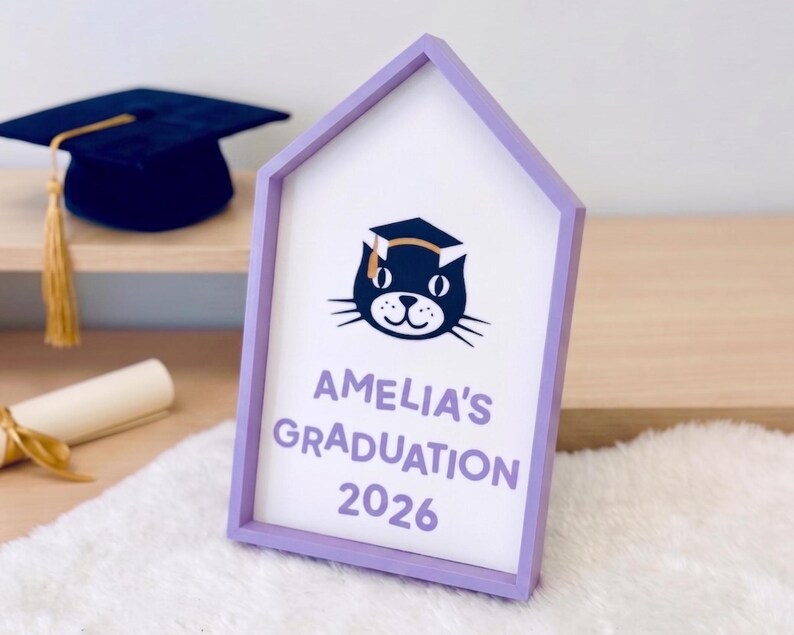 May include: A house-shaped sign with a lavender frame, featuring a cat wearing a graduation cap. The sign reads "AMELIA'S GRADUATION 2026" in lavender lettering. A graduation cap and diploma are visible in the background.