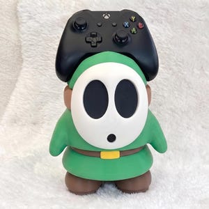 May include: A green and brown character figure with a white oval mask featuring two large black eye holes and a small black mouth. A black wireless game controller is positioned on top of the figure's head, resting between its ears.