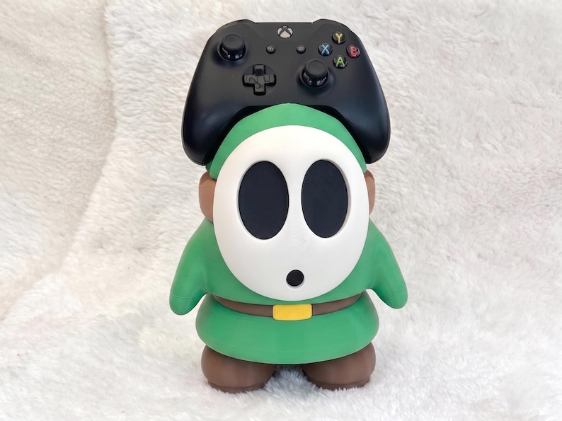 May include: A green and brown character figure with a white oval mask featuring two large black eye holes and a small black mouth. A black wireless game controller is positioned on top of the figure's head, resting between its ears.