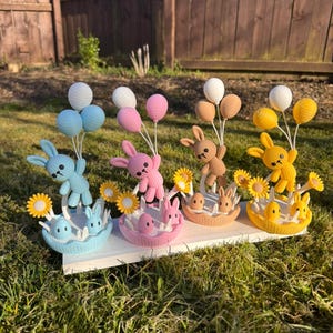 May include: A display of four Easter-themed decorations. Each features a crocheted bunny holding balloons, with a basket containing eggs and smaller bunnies. The color scheme includes blue, pink, brown, and yellow, with yellow sunflowers.