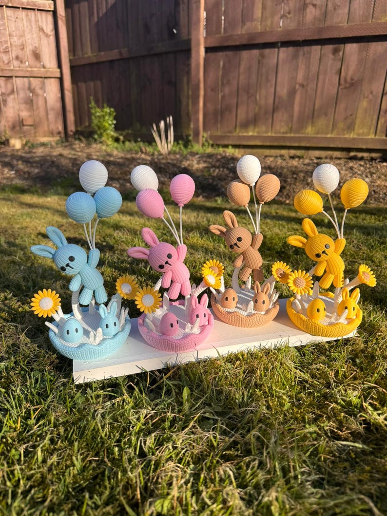 May include: A display of four Easter-themed decorations. Each features a crocheted bunny holding balloons, with a basket containing eggs and smaller bunnies. The color scheme includes blue, pink, brown, and yellow, with yellow sunflowers.