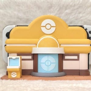 May include: A yellow and white decorative stand designed to hold a gaming console. The stand is shaped like a building with a circular logo on the front. The stand has a brown base and a light blue center.