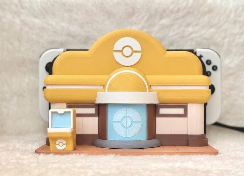 May include: A yellow and white decorative stand designed to hold a gaming console. The stand is shaped like a building with a circular logo on the front. The stand has a brown base and a light blue center.