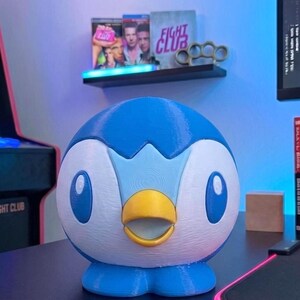 K&ouml;nnte beinhalten: Eine 3D-gedruckte Piplup-Figur, ein blau-wei&szlig;es pinguinartiges Pok&eacute;mon, steht auf einer schwarzen Oberfl&auml;che. Die Figur hat einen gelben Schnabel und blaue Augen. Im Hintergrund zeigt ein Regal Filmh&uuml;llen und Schlagringe.