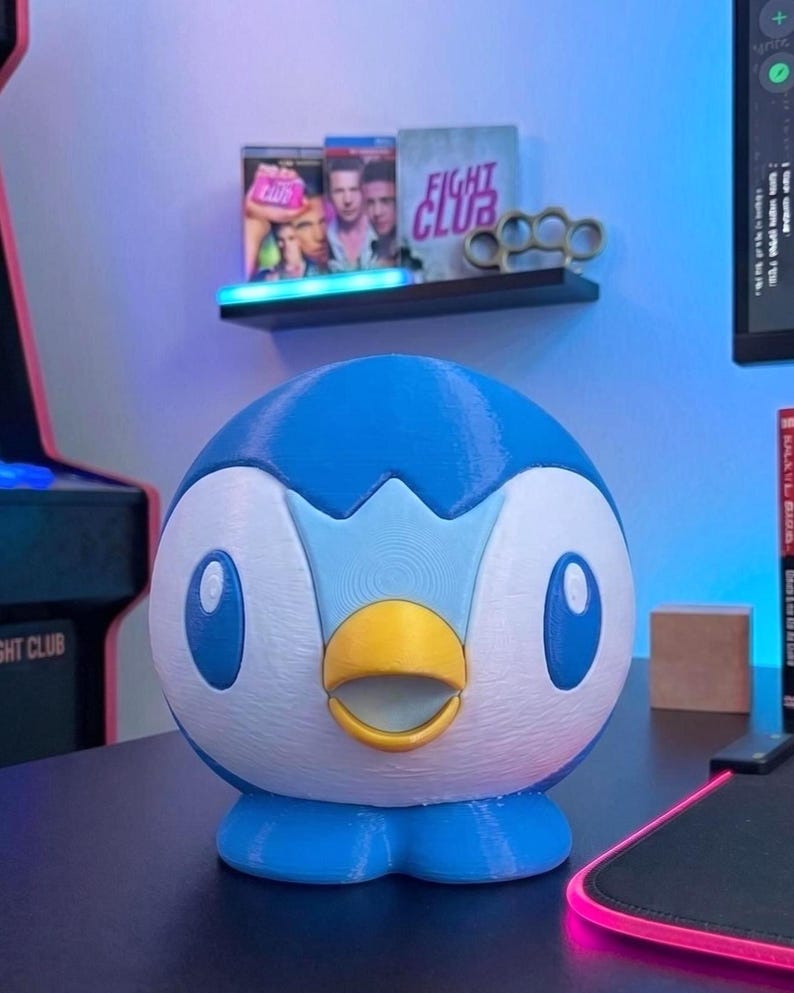 K&ouml;nnte beinhalten: Eine 3D-gedruckte Piplup-Figur, ein blau-wei&szlig;es pinguinartiges Pok&eacute;mon, steht auf einer schwarzen Oberfl&auml;che. Die Figur hat einen gelben Schnabel und blaue Augen. Im Hintergrund zeigt ein Regal Filmh&uuml;llen und Schlagringe.