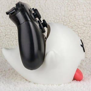 May include: A white ghost-shaped controller stand with a smooth matte finish, featuring a small red tongue protruding from the side and black facial features, currently holding a black gaming controller upright in its open top section.