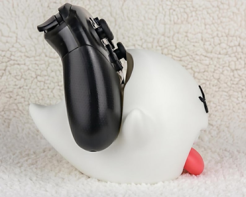 May include: A white ghost-shaped controller stand with a smooth matte finish, featuring a small red tongue protruding from the side and black facial features, currently holding a black gaming controller upright in its open top section.