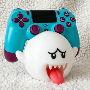 May include: A bright turquoise and dark purple video game controller resting on a white, spherical ghost-shaped stand. The stand features a wide, open mouth with sharp white teeth and a protruding red tongue, with black eyes and arched eyebrows.