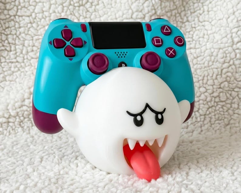 May include: A bright turquoise and dark purple video game controller resting on a white, spherical ghost-shaped stand. The stand features a wide, open mouth with sharp white teeth and a protruding red tongue, with black eyes and arched eyebrows.