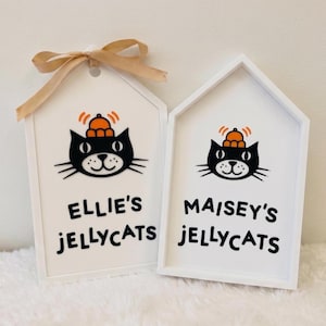 Puede incluir: Dos letreros de madera blancos en forma de casa con una cinta beige atada al de la izquierda. Cada letrero presenta una ilustraci&oacute;n de un gato negro con un sombrero naranja y el texto negro Ellie's JELLYCATS a la izquierda y Maisey's JELLYCATS a la derecha.