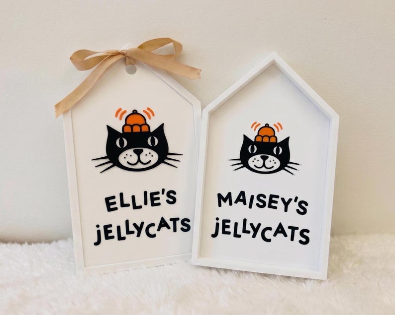 Puede incluir: Dos letreros de madera blancos en forma de casa con una cinta beige atada al de la izquierda. Cada letrero presenta una ilustraci&oacute;n de un gato negro con un sombrero naranja y el texto negro Ellie's JELLYCATS a la izquierda y Maisey's JELLYCATS a la derecha.