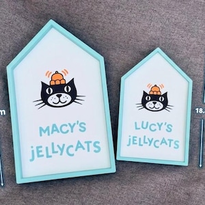 May include: Two house-shaped signs with a light blue border. The signs feature a black cat illustration wearing a hat, and the text "MACY'S JELLYCATS" and "LUCY'S JELLYCATS". The larger sign is 26.2 cm tall, and the smaller sign is 18.8 cm tall.