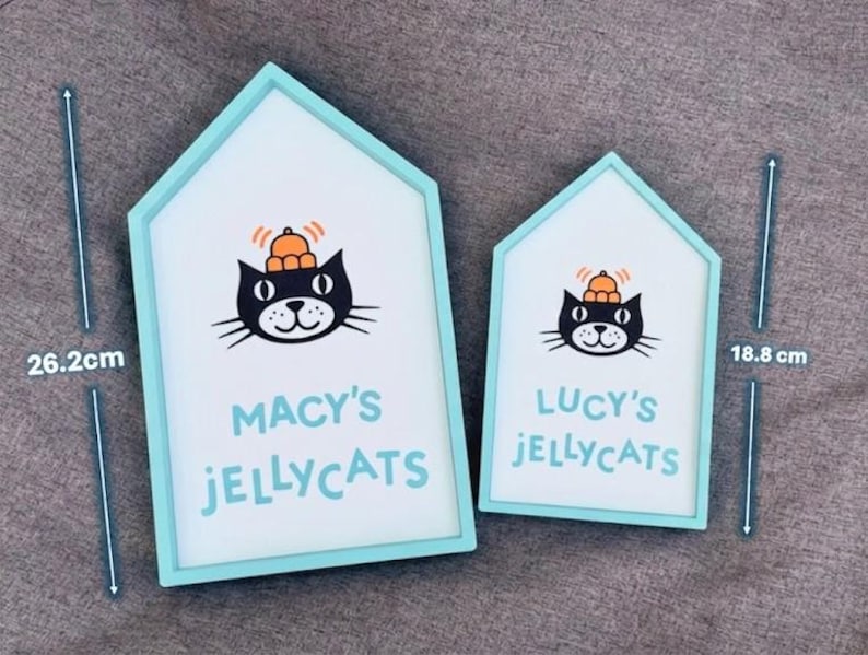 May include: Two house-shaped signs with a light blue border. The signs feature a black cat illustration wearing a hat, and the text "MACY'S JELLYCATS" and "LUCY'S JELLYCATS". The larger sign is 26.2 cm tall, and the smaller sign is 18.8 cm tall.