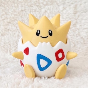 May include: A yellow and white figurine of a creature emerging from a cracked egg shell. The creature has a star-shaped head with black eyes and a smile. The white egg shell features red and blue triangular patterns on its rounded surface.
