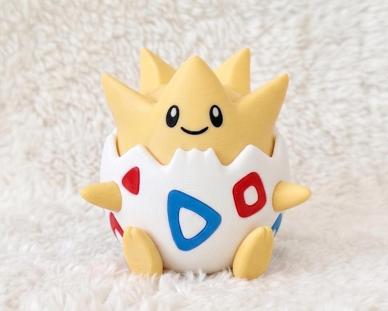 May include: A yellow and white figurine of a creature emerging from a cracked egg shell. The creature has a star-shaped head with black eyes and a smile. The white egg shell features red and blue triangular patterns on its rounded surface.