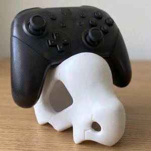 May include: A black Nintendo Switch Pro controller rests on a white, skull-shaped stand. The controller has black buttons and joysticks. The stand is a decorative item, likely 3D-printed, and is designed to hold the controller.