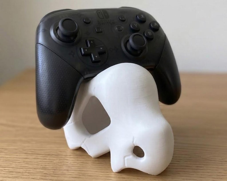 May include: A black Nintendo Switch Pro controller rests on a white, skull-shaped stand. The controller has black buttons and joysticks. The stand is a decorative item, likely 3D-printed, and is designed to hold the controller.