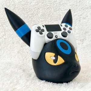 May include: A black and blue gaming controller stand shaped like a stylized creature head with yellow eyes and pointed ears. A white wireless game controller with black buttons and joysticks is resting securely on top of the stand.