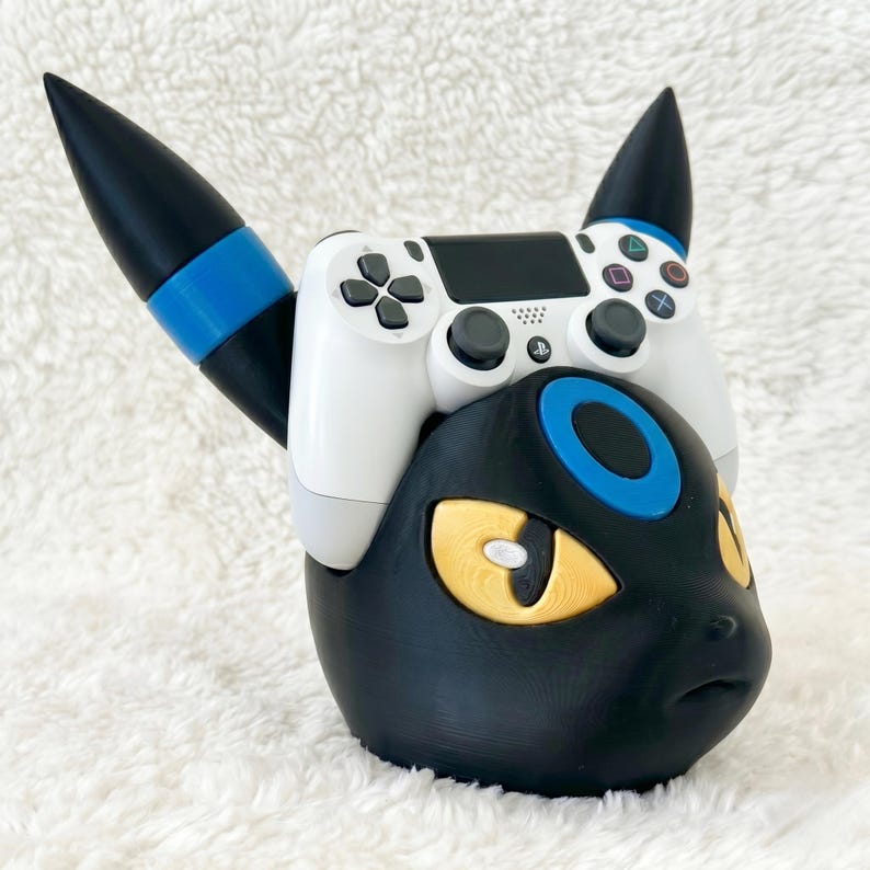May include: A black and blue gaming controller stand shaped like a stylized creature head with yellow eyes and pointed ears. A white wireless game controller with black buttons and joysticks is resting securely on top of the stand.