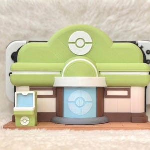 May include: A 3D-printed model of a building with a green roof and white accents, resembling a video game structure. The model has a light green and brown color scheme, with a light blue circular design on the front. A video game console is visible behind the model.