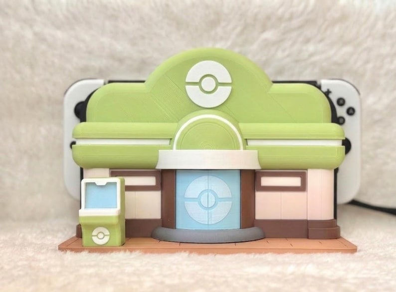 May include: A 3D-printed model of a building with a green roof and white accents, resembling a video game structure. The model has a light green and brown color scheme, with a light blue circular design on the front. A video game console is visible behind the model.