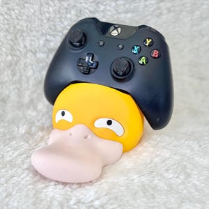 Puede incluir: Un mando de videojuegos inal&aacute;mbrico negro apoyado sobre un soporte con forma de pato amarillo y rosa claro. El mando cuenta con una cruceta, dos joysticks anal&oacute;gicos y cuatro botones de acci&oacute;n de colores etiquetados como A, B, X e Y, con un logo Xbox.