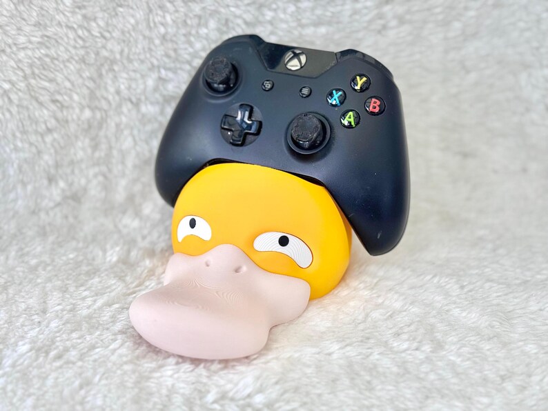 Puede incluir: Un mando de videojuegos inal&aacute;mbrico negro apoyado sobre un soporte con forma de pato amarillo y rosa claro. El mando cuenta con una cruceta, dos joysticks anal&oacute;gicos y cuatro botones de acci&oacute;n de colores etiquetados como A, B, X e Y, con un logo Xbox.