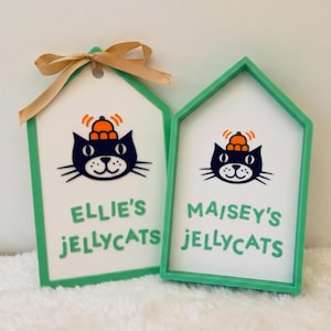 Puede incluir: Dos letreros en forma de casa con marcos verde menta y centros blancos. Cada uno presenta una ilustraci&oacute;n de un gato negro con un sombrero naranja y el texto "ELLIE'S JELLYCATS" y "MAISEY'S JELLYCATS" en verde. El letrero izquierdo tiene un lazo beige.