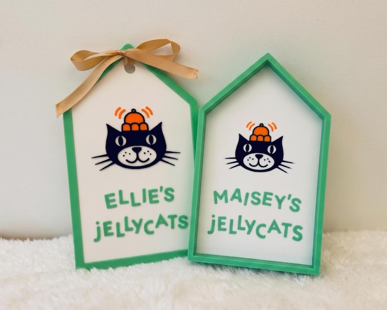 Puede incluir: Dos letreros en forma de casa con marcos verde menta y centros blancos. Cada uno presenta una ilustraci&oacute;n de un gato negro con un sombrero naranja y el texto "ELLIE'S JELLYCATS" y "MAISEY'S JELLYCATS" en verde. El letrero izquierdo tiene un lazo beige.